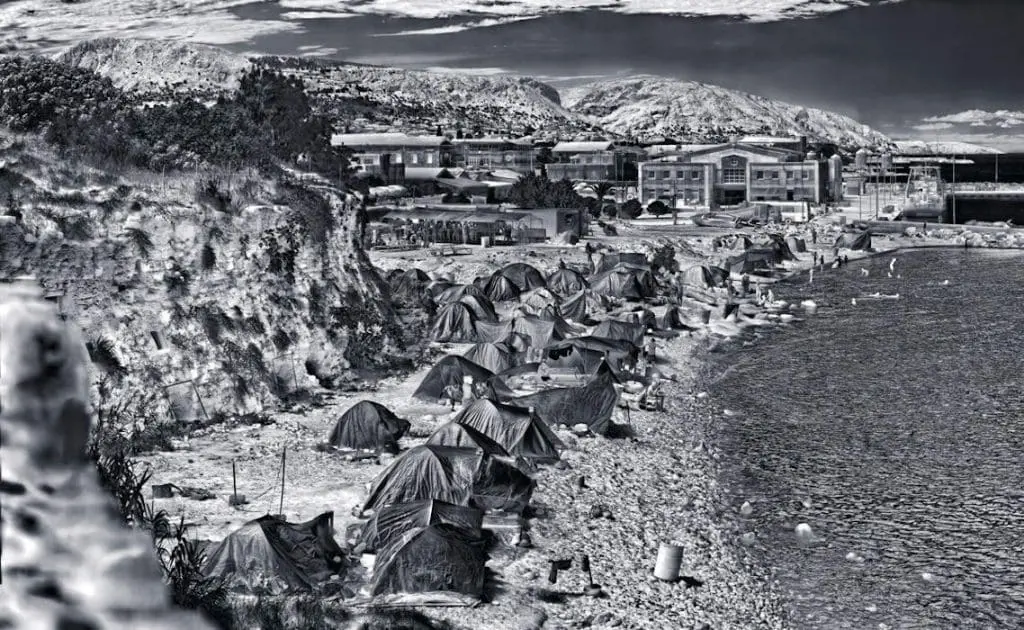 © Richard Mosse Souda Camp, Chios Island, Greece, 2017 ** MOCAK Collection, Krakow Situato ai piedi dei bastioni di una Fortezza medievale, Souda ospita 950 persone secondo le stime ufficiali, una cifra che corrisponde al doppio della capienza, con i nuovi arrivati costretti a montare le tende sulla spiaggia adiacente. I residenti lamentano infestazioni di ratti, epidemie di scabbia, scarsità di alloggi e intossicazioni alimentari. Il 30% dei richiedenti asilo di Souda hanno trascorso nel campo più di sei mesi. Gli abitanti dell’isola di Chios, furiosi per il fatto che la loro terra e la sua florida industria turistica siano in prima linea nella crisi europea dei migranti, organizzano frequenti proteste. Il partito neofascista Alba Dorata raccoglie nell’isola numerosi consensi e i suoi membri hanno attaccato il campo in varie occasioni, lanciando dalle mura del castello massi e bombe molotov sui migranti indifesi parecchi metri più in basso e ferendo molte persone. I rifugiati sono spesso aggrediti e incarcerati illegalmente da poliziotti che simpatizzano con gli ideali di Alba Dorata. Sono stati riportati casi di violenza sessuale ai danni di giovani donne ad opera di cittadini di Chios, documentati da video girati con il cellulare e diffusi a scopo di intimidazione. Secondo Human Rights Watch, i casi di attacchi d’ansia, autolesionismo e suicidio tra i profughi di Chios sono molto numerosi.